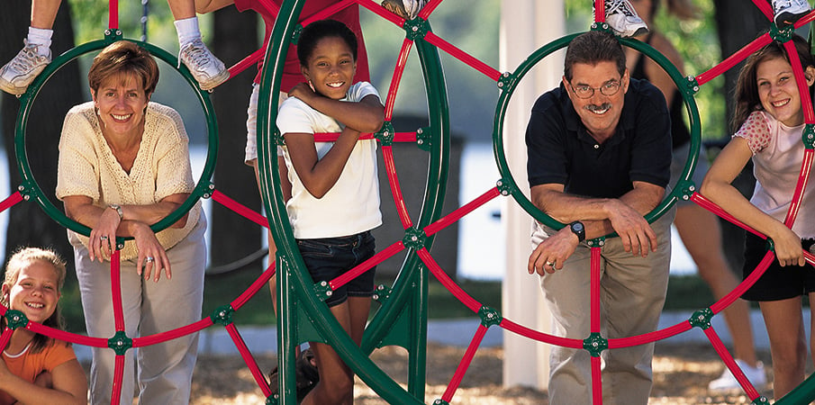 Landscape Structure founders Steve and Barb King and a group of children smiling leaning through a Spider Web Climber. 