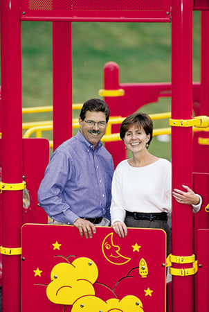Landscape Structure founders Steve and Barb King smile while standing behind a Permaline panel on a playground. 