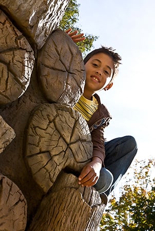 A boy smiling at camera while leaning over a  log stack climber with wood grain hand holds.