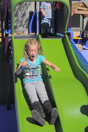 A girl with her mouth open slides down a green slide. 
