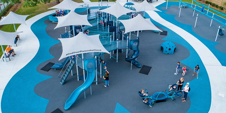 Overhead view of a light blue and dark blue playground with numerous blue bridges between different playground structures, covered by light blue shade structures, one with the word "#FindYourPlaceintheCloud" on the shade. 