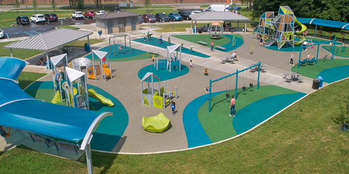 A sunny day overlooking a blue, green and light brown playground with two Alpha Towers in the background with a green slide and bright green, silver and turquoise playground components in the foreground. 