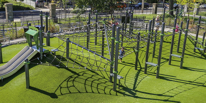 Grey climbing structure with children playing behind it in the distance against green turf surfacing. 