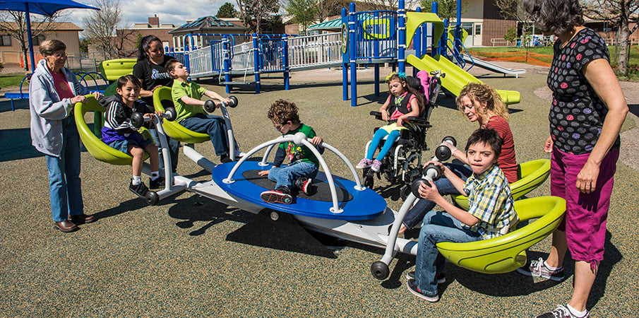 Children play on the inclusive We-saw seesaw as three women stand by watching.
