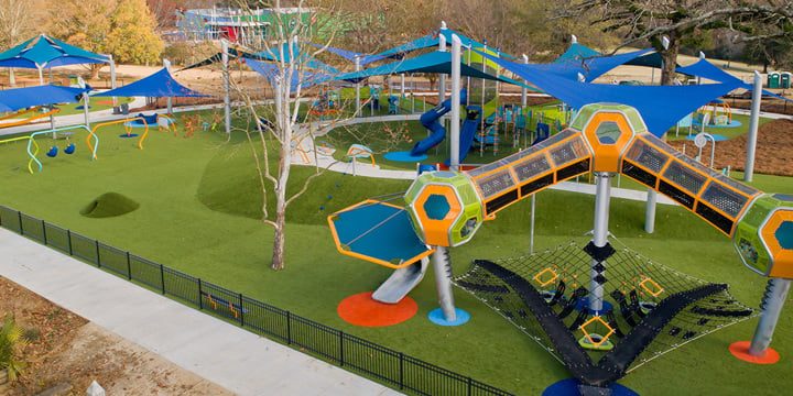 Tall geometric playground tower in orange and bright green with black net structure beneath  it with more playground components in the background and royal blue SkyWays shade structures. 