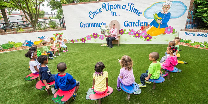 Children sit in a circle facing a sign that reads "Once Upon a Time in... Grammy's Garden" while a woman reads a book to them. 