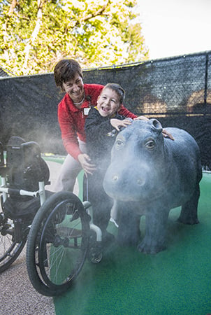A woman and a boy stand next to a hippo sculpture and the boy laughs as water sprays out of the hippo's nose. There is a wheelchair to the side. 