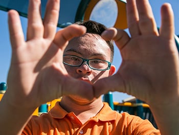 A boy makes a circle with his fingers and puts his hands up to the camera lens so that his face is framed.